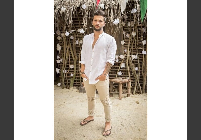 homem com camisa branca dobrada, calça bege e chinelo em frente a uma cabana de praia
