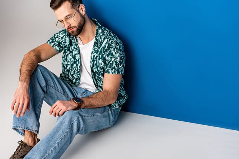 modelo sentado escorado em uma parede azul vestindo calça jeans, camisa florida aberta e uma camiseta branca