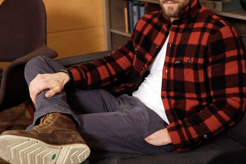 homem sentado em uma cadeira utilizando blusa xadrez, calça jeans e bota